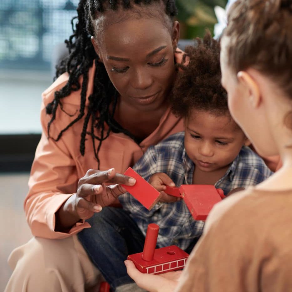 Young black woman and her wife explaining little son how to build house out of colorful wooden blocks